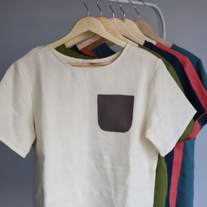 Pu&ograve; includere: Una t-shirt di lino bianco con una tasca a toppa marrone. La t-shirt &egrave; appesa a un appendiabiti di legno con altre t-shirt di vari colori.