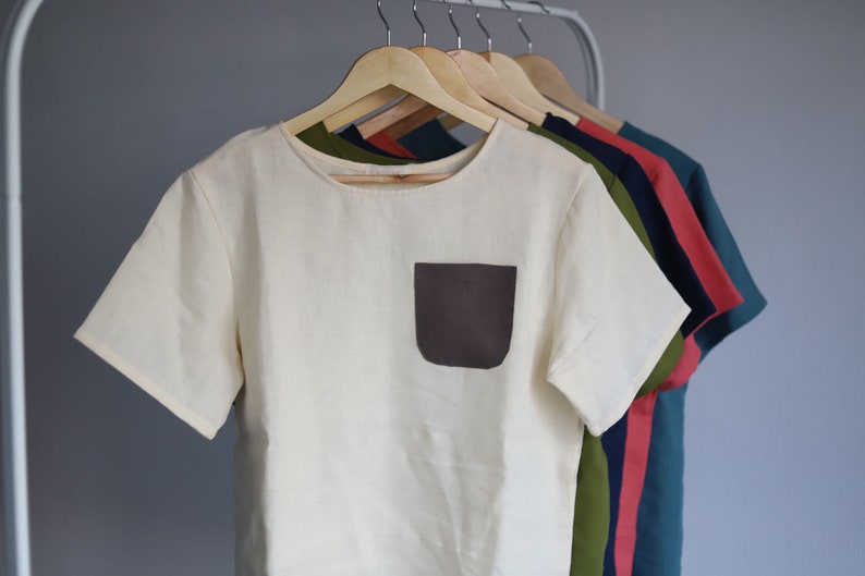 Pu&ograve; includere: Una t-shirt di lino bianco con una tasca a toppa marrone. La t-shirt &egrave; appesa a un appendiabiti di legno con altre t-shirt di vari colori.