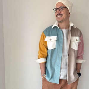 May include: A man wearing a white beanie, black framed glasses, and a multi-colored corduroy shirt with a white t-shirt underneath. He is wearing brown corduroy pants and has a watch on his left wrist.