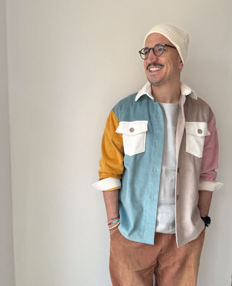 May include: A man wearing a white beanie, black framed glasses, and a multi-colored corduroy shirt with a white t-shirt underneath. He is wearing brown corduroy pants and has a watch on his left wrist.