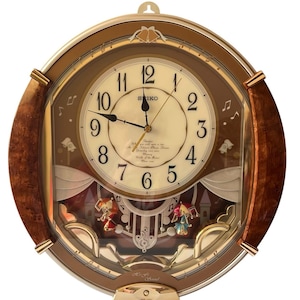May include: A round Seiko wall clock with a wood-grain frame, a cream-colored face with black Arabic numerals, and gold-tone hands. The lower section features two small figures, musical notes, and a castle graphic behind a clear glass panel.