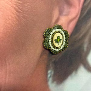 May include: A floral-shaped earring with a central oval design featuring a green frog illustration. The earring has a cream-colored background, surrounded by white beads, green lace, and gold-colored edges. The earring is a unique accessory.
