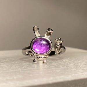 May include: A silver-toned ring featuring a bunny design with a purple gemstone center. The bunny has upright ears and a carrot detail on the side. The ring is set on a white surface.