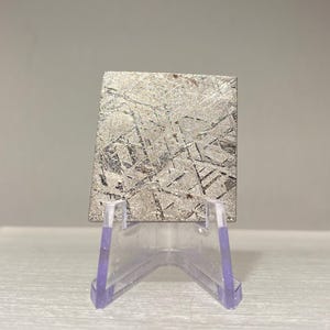 Puede incluir: Un espécimen de meteorito rectangular plateado con un patrón de Widmanstätten único, exhibido en un soporte de acrílico transparente. La superficie del meteorito presenta líneas entrecruzadas y estructuras cristalinas.