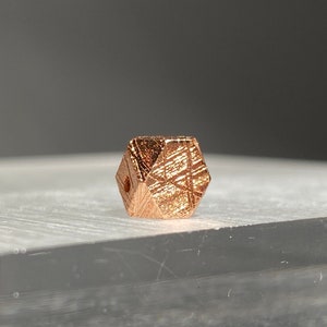 May include: A small, geometric copper-colored bead with a faceted design. The bead has a series of intersecting lines etched across its surface, creating a textured appearance. The bead is displayed on a clear, rectangular surface.