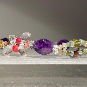 May include: A beaded bracelet featuring a large, faceted amethyst stone and a variety of colorful beads. The bracelet includes clear, pink, yellow, and green beads, with a silver clasp. The design is delicate and intricate.