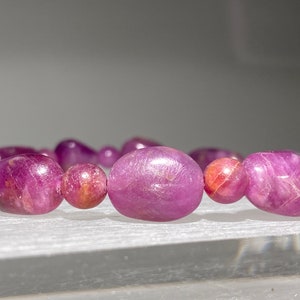 May include: A close-up of a pink gemstone bracelet with oval-shaped beads. The beads are smooth and have a slight shimmer.
