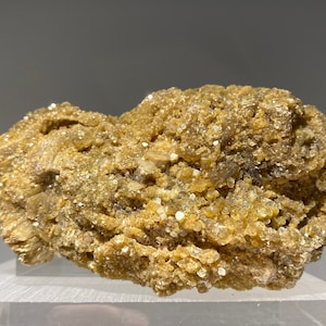 May include: A cluster of small, golden-brown crystals on a rough, grey rock. The crystals are tightly packed together, creating a textured surface.