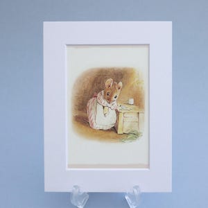 Pode incluir: Ilustração emoldurada de um rato usando um vestido rosa e avental, em pé perto de uma pequena mesa com uma xícara. A obra de arte tem uma estética vintage, com cores suaves e um estilo detalhado. A moldura é branca.