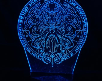 May include: A blue LED night light with a circular design featuring a stylized depiction of Cthulhu, a fictional cosmic entity from the works of H.P. Lovecraft. The design includes a pentagram, a crescent moon, and a circle of text in a gothic font.