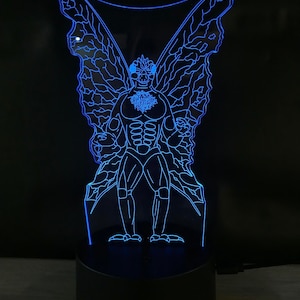 May include: A blue LED night light with a clear acrylic panel depicting a winged creature with a human-like body. The creature has a large head with a pointed snout and large eyes. The wings are spread wide and have a detailed vein pattern.
