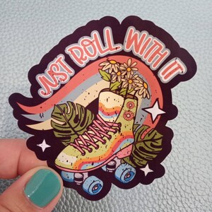 May include: A colourful sticker featuring a pair of roller skates with flowers inside. The text "Just Roll With It" is written in a retro font.