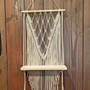 May include: A macrame wall hanging with a wooden shelf. The macrame is made with natural colored cord and has a diamond pattern. The shelf is made of light wood and is attached to the bottom of the macrame.