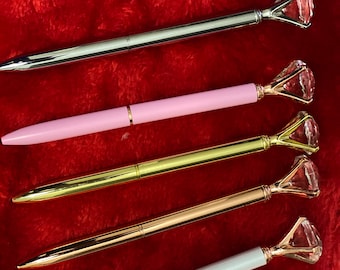 May include: Five pens with diamond-shaped tops in different colors: silver, pink, gold, rose gold, and white. The pens are on a red fuzzy surface.