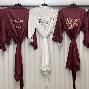 Peut inclure: Cinq robes en satin avec bordure en dentelle, suspendues à des cintres. Les robes sont bordeaux, sauf une qui est blanche. Les robes portent des inscriptions dorées qui disent "Bride", "Bridesmaid", "Maid of Honor", "Flower Girl", et "Bridesmaid".