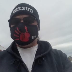 May include: A person wearing a black hooded sweatshirt, a black beanie with the word "GUZZI" embroidered on it, and a black face mask with a red handprint and the word "WIMM" printed on it.