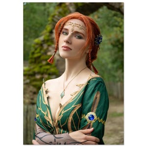 Peut inclure: Une femme aux cheveux roux et aux yeux verts, vêtue d'une robe verte avec des détails dorés et un diadème doré. La robe présente un décolleté plongeant et des ornements bleus. L'arrière-plan est une scène extérieure floue.