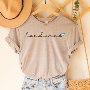 May include: A gray t-shirt with the word "honduras" written in black cursive with a blue and white heart featuring the Honduran flag. The shirt is hanging on a wooden rack with a straw hat and a woven bag.