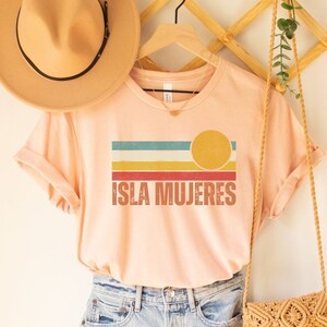 May include: A pink t-shirt with a retro-style graphic of a sunset and the text "ISLA MUJERES". The shirt is hanging on a wooden hanger with a straw hat and a woven bag.