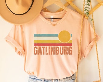 May include: A peach-colored t-shirt with a retro-style graphic of a sun and stripes in red, yellow, blue, and green. The text "GATLINBURG" is printed in brown below the graphic.