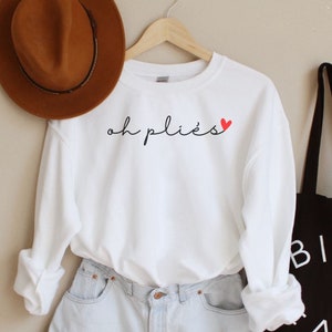 May include: A white sweatshirt with the text "oh pliés" in black script and a red heart. The sweatshirt is paired with light wash denim shorts, a brown hat, and a black tote bag with white text that reads "BIEN FAIT".