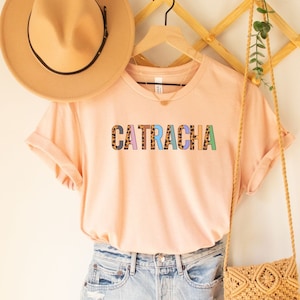 May include: A light pink t-shirt with the word "CATRACHA" printed in colorful letters with a leopard print pattern. The shirt is hanging on a wooden hanger with a brown hat and a woven bag.