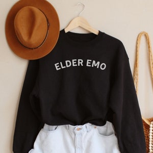 May include: Black sweatshirt with the text "ELDER EMO" in white lettering, white denim shorts, a brown fedora hat, and a crocheted bag with white beads.