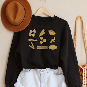 Op de afbeelding: Zwart sweatshirt met een afbeelding van verschillende soorten pasta in goud. Het sweatshirt wordt gedragen met lichtblauwe denim shorts en een bruine hoed.