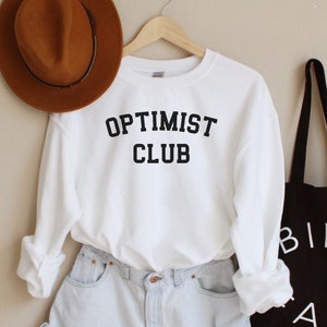 May include: White sweatshirt with the text "OPTIMIST CLUB" in black lettering. The sweatshirt is paired with light blue denim shorts and a brown fedora. A black tote bag with the text "BIEN FAIT" in white lettering is also pictured.