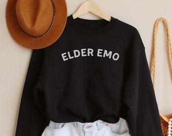 May include: Black sweatshirt with the text "ELDER EMO" in white lettering, white denim shorts, a brown fedora hat, and a crocheted bag with white beads.