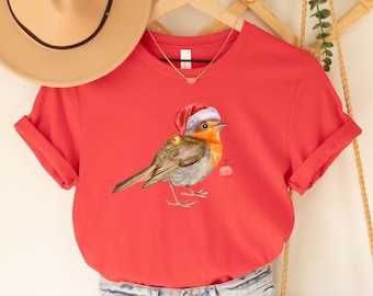 May include: Red t-shirt with a robin wearing a Santa hat and holding a gift. The shirt is on a hanger with a straw hat and a woven bag.