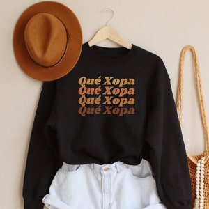 May include: Black sweatshirt with the text "Qué Xopa" repeated four times in a retro font with orange, brown, and yellow colors. The sweatshirt is worn with light blue denim shorts and a brown fedora.