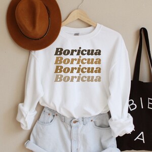 May include: White long-sleeved sweatshirt with the word "Boricua" repeated four times in brown, tan, and dark brown lettering. The sweatshirt is worn with light blue denim shorts and a brown fedora hat. A black tote bag with the word "BIEN AIT" in white lettering is also pictured.