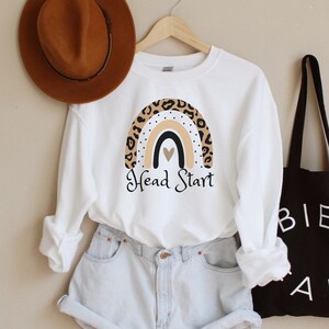 May include: White sweatshirt with a rainbow graphic and the text "Head Start". The rainbow is decorated with a leopard print pattern. The sweatshirt is paired with light blue denim shorts and a brown fedora.