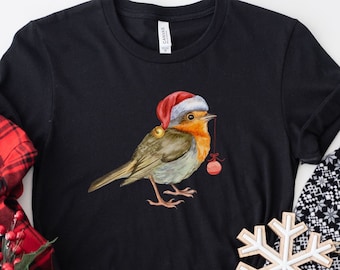 May include: Black t-shirt featuring a watercolor illustration of a robin wearing a Santa hat and holding a Christmas ornament. The bird has orange, brown, and gray feathers. A red and black plaid shirt and snowflake decoration are also visible.