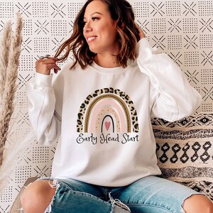 May include: A white sweatshirt with a rainbow design and the text "Early Head Start". The rainbow is made of pink, gold, and brown stripes with a small pink heart in the center. The rainbow is surrounded by a leopard print pattern.