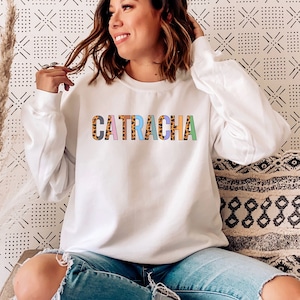 May include: A white sweatshirt with the word "CATRACHA" printed in colorful letters with a leopard print design. The model is wearing blue jeans and sitting on a couch.