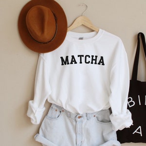 May include: White sweatshirt with the word "MATCHA" printed in black. The sweatshirt is paired with light blue denim shorts and a black tote bag with the words "BIEN FAIT" printed in white.
