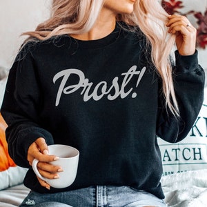 May include: A black sweatshirt with the word "Prost!" written in white cursive lettering. The sweatshirt is being worn by a person sitting on a couch.