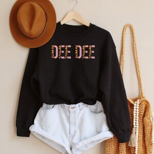 May include: Black sweatshirt with the text "DEE DEE" in pink letters with a leopard print pattern. The sweatshirt is worn with light blue denim shorts and a brown woven bag with white beads.