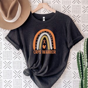 May include: A dark grey heather t-shirt featuring a rainbow graphic and the text "CRPS WARRIOR". The rainbow has a heart and an orange ribbon in the centre.