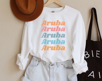 May include: White long-sleeve sweatshirt with the word "Aruba" repeated five times in a rainbow color pattern. The sweatshirt is paired with denim shorts and a black tote bag with the text "BIEN AIT" in white.