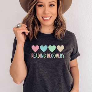 May include: A dark grey t-shirt with a row of four pastel colored hearts and the text "READING RECOVERY".