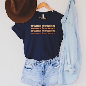 May include: A navy blue t-shirt with the text "women in science" repeated three times in a gradient of orange, yellow, and red. The shirt is on a hanger and is styled with a brown hat, denim shorts, and a light blue denim jacket.