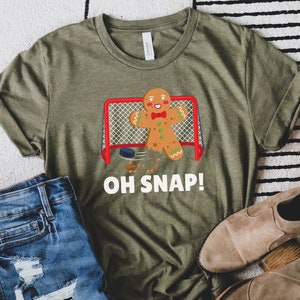 May include: A green t-shirt with a gingerbread man playing hockey. The gingerbread man is wearing a red bow tie and is holding a hockey stick. The text "OH SNAP!" is printed below the image.