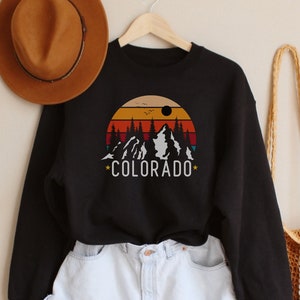 May include: Black sweatshirt with a retro mountain scene and the text "*COLORADO*" printed on the front. The sweatshirt is paired with light blue denim shorts and a brown woven bag.
