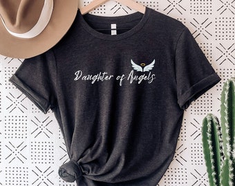 May include: Dark grey heather t-shirt with the text "Daughter of Angels" and a white angel wing graphic. The shirt is hanging on a wooden hanger against a white background with a black and white geometric pattern.