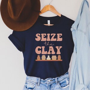 May include: A navy blue t-shirt with the text "Seize the Clay" in pink and white lettering. The shirt has a graphic of five clay pots in different shades of brown on a shelf.