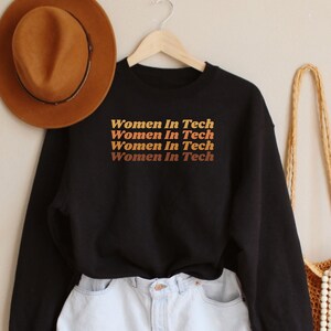 Op de afbeelding: Zwart sweatshirt met de tekst "Women in Tech" vier keer herhaald in een verloop van oranje, geel en rood. De tekst is in een retro-lettertype stijl.