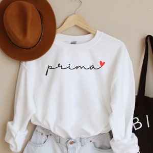 May include: White sweatshirt with the word "prima" in black script and a small red heart. The sweatshirt is worn with light blue denim shorts and a brown fedora.